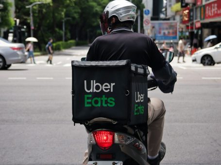A delivery rider on a motorcycle with an Uber Eats bag waits at a city intersection.
