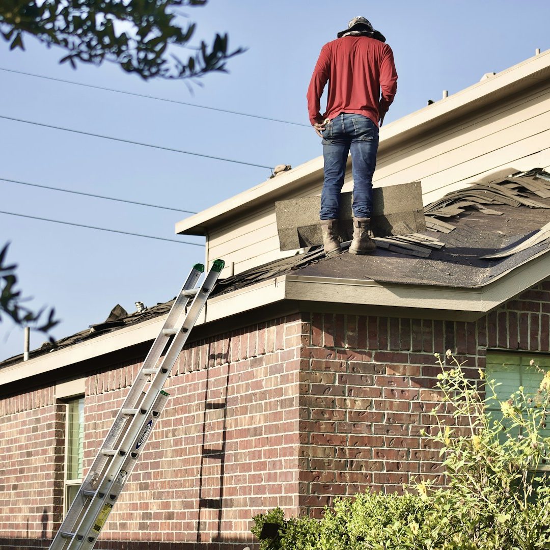 A person in a red sweater stands on a roof, repairing shingles near a ladder.