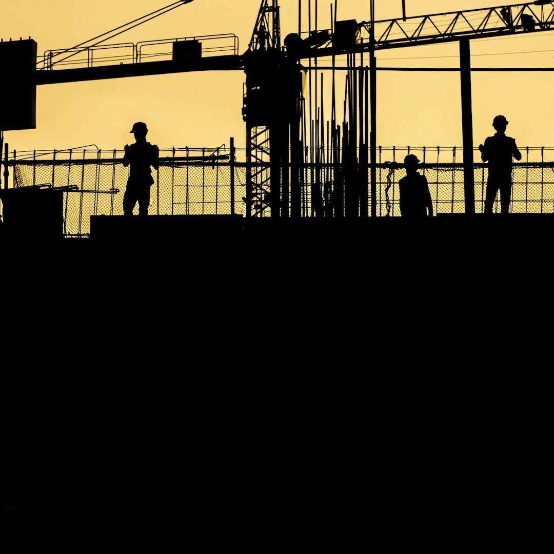 Silhouetted construction workers against a yellow sunset with cranes in the background.