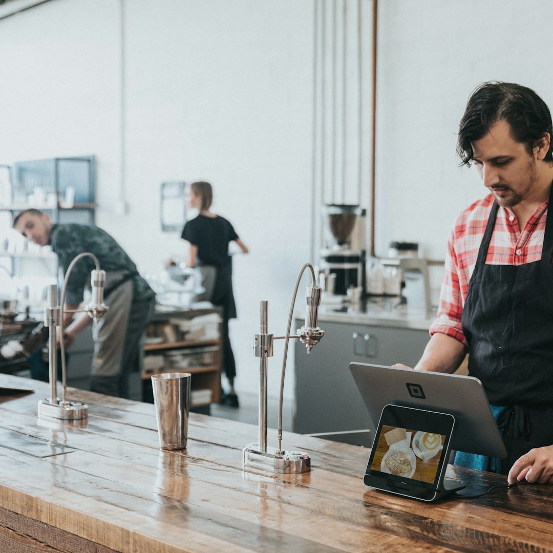 Barista at a counter using a tablet, with another worker preparing coffee in the background.