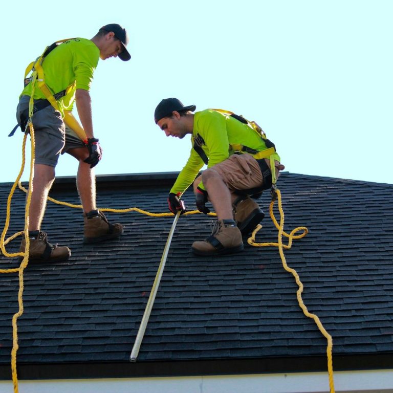 Two workers in bright shirts measure a roof while secured with ropes.